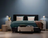 Et moderne soveværelse med en seng, der har et mørkt polstret hovedgærde, teal sengetøj og to store, bløde hvide puder. Sengen er flankeret af to hvide natborde, hver med en moderne lampe og dekorative genstande som en vase med gule blomster og en bakke med en karaffel. For enden af sengen står en tekstureret, lysefarvet bænk med sammenfoldede tæpper og en pude ovenpå. Rummet har et minimalistisk design med en blå accentvæg og lyst trægulv.