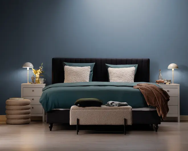 Et moderne soveværelse med en seng, der har et mørkt polstret hovedgærde, teal sengetøj og to store, bløde hvide puder. Sengen er flankeret af to hvide natborde, hver med en moderne lampe og dekorative genstande som en vase med gule blomster og en bakke med en karaffel. For enden af sengen står en tekstureret, lysefarvet bænk med sammenfoldede tæpper og en pude ovenpå. Rummet har et minimalistisk design med en blå accentvæg og lyst trægulv.