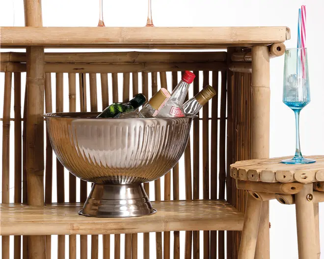 Stor, rund metalskål i sølvfarve med riflede sider og en solid fod. Skålen er fyldt med is og flere flasker drikkevarer, herunder klare og grønne glasflasker med forskellige farvede kapsler. Skålen står på en bambusreol, hvilket giver et rustikt og naturligt udtryk. Til højre står et højt, blåtonet glas med lang stilk og to farverige sugerør på en tilsvarende bambusoverflade.