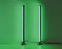 Et par slanke, lodrette LED-lysstænger med moderne design, der udsender et kraftigt grønt skær. Hver lysstang står på en rund, solid sort fod og er forbundet med en synlig strømkabel. Lysene er placeret tæt på en væg, hvilket giver en jævn og intens grøn belysning på overfladen bag dem.