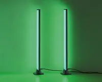 Et par slanke, lodrette LED-lysstænger med moderne design, der udsender et kraftigt grønt skær. Hver lysstang står på en rund, solid sort fod og er forbundet med en synlig strømkabel. Lysene er placeret tæt på en væg, hvilket giver en jævn og intens grøn belysning på overfladen bag dem.