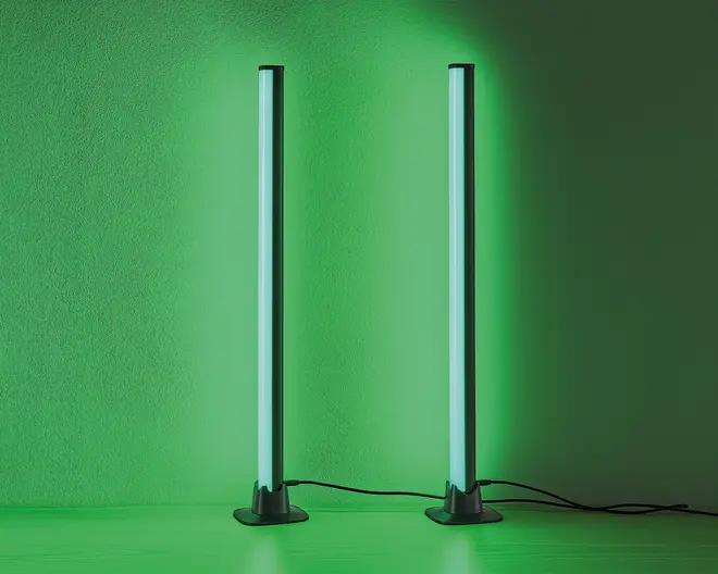 Et par slanke, lodrette LED-lysstænger med moderne design, der udsender et kraftigt grønt skær. Hver lysstang står på en rund, solid sort fod og er forbundet med en synlig strømkabel. Lysene er placeret tæt på en væg, hvilket giver en jævn og intens grøn belysning på overfladen bag dem.