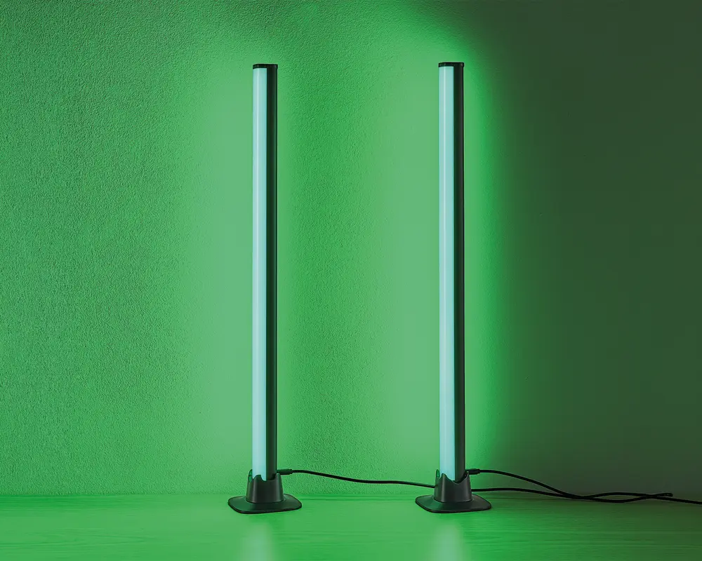 Et par slanke, lodrette LED-lysstænger med moderne design, der udsender et kraftigt grønt skær. Hver lysstang står på en rund, solid sort fod og er forbundet med en synlig strømkabel. Lysene er placeret tæt på en væg, hvilket giver en jævn og intens grøn belysning på overfladen bag dem.