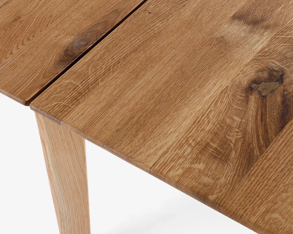 Nærbillede af en træoverflade med naturlig finish, hvor åremønster, knaster og en sprække mellem to planker er tydelige. Træet har en varm, mellembrun farve og en mat tekstur. Benet er lavet af samme træ med tilsvarende åremønster.