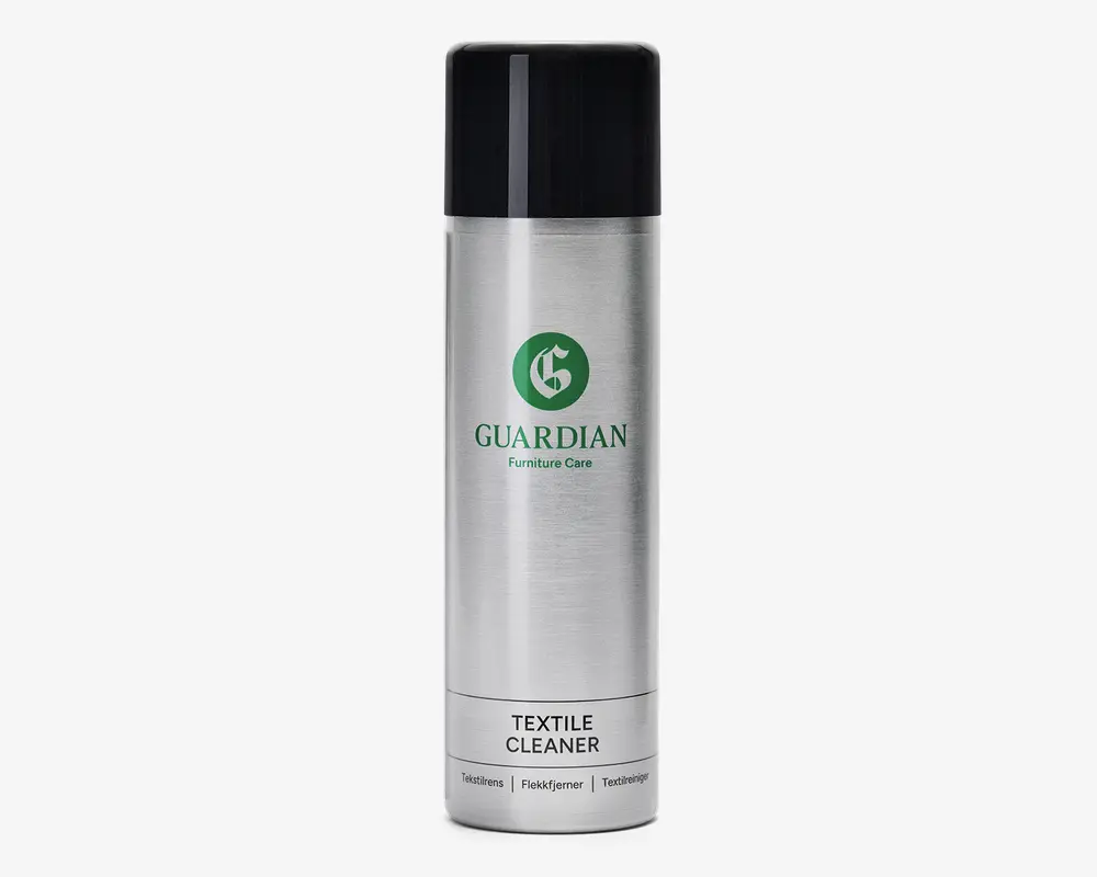 En høj, cylindrisk spraydåse med sølvfarvet krop og sort låg. Forsiden har et grønt logo og teksten 'Guardian Furniture Care' i grønt, samt 'Textile Cleaner' trykt med sort på en hvid etiket nær bunden. Dåsen har et stilrent, moderne design og ser nem ud at holde og bruge.