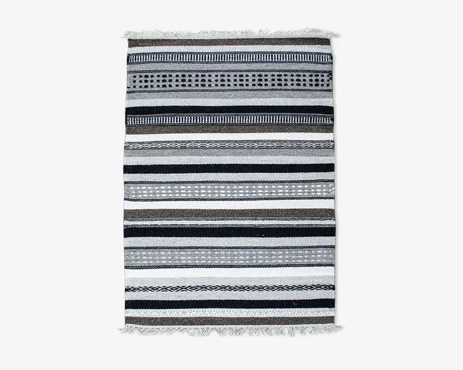 Rektangulært vævet tekstil med vandrette striber i sorte, hvide og grå nuancer. Striberne varierer i bredde og har forskellige mønstre som prikker, diamanter og ensfarvede linjer. Begge ender af tekstilet har korte, knyttede frynser.