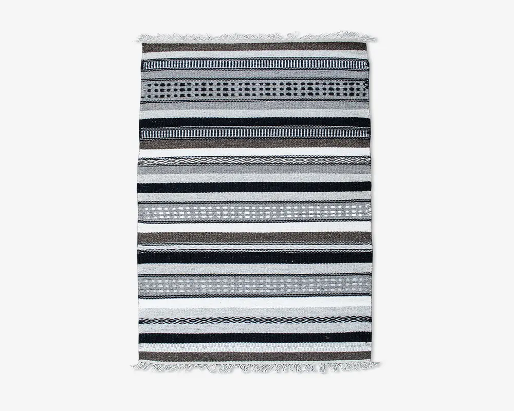 Rektangulært vævet tekstil med vandrette striber i sorte, hvide og grå nuancer. Striberne varierer i bredde og har forskellige mønstre som prikker, diamanter og ensfarvede linjer. Begge ender af tekstilet har korte, knyttede frynser.