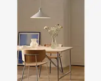 Et moderne spiseområde med et rektangulært bord med lys træbordplade og ben af metal. På bordet står en klar glasvase med en buket vilde blomster, en hvid keramikkande og flere klare drikkeglas. To stole med buede træryglæn og metalstel står ved bordet. I baggrunden ses en hvid murstensvæg, et stort indrammet blå-hvidt kunstværk, der står på gulvet, samt en minimalistisk hvid pendellampe, der hænger over bordet. Stilen er enkel og moderne med naturlige og neutrale farver.