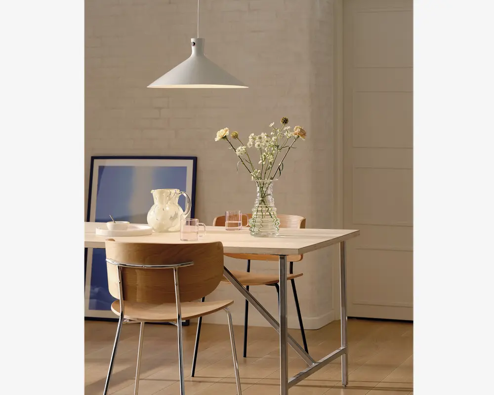 Et moderne spiseområde med et rektangulært bord med lys træbordplade og ben af metal. På bordet står en klar glasvase med en buket vilde blomster, en hvid keramikkande og flere klare drikkeglas. To stole med buede træryglæn og metalstel står ved bordet. I baggrunden ses en hvid murstensvæg, et stort indrammet blå-hvidt kunstværk, der står på gulvet, samt en minimalistisk hvid pendellampe, der hænger over bordet. Stilen er enkel og moderne med naturlige og neutrale farver.