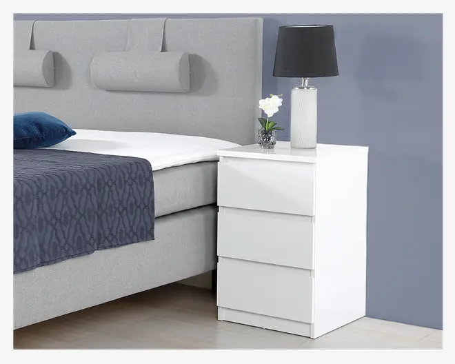 Et moderne, hvidt sengebord med blank overflade og tre skuffer uden greb. Skufferne har et minimalistisk, fladt frontdesign. Bordet er rektangulært med rene, lige kanter. Ovenpå står en hvid lampe med rillet struktur og sort lampeskærm samt en lille dekorativ plante i en glaspotte.