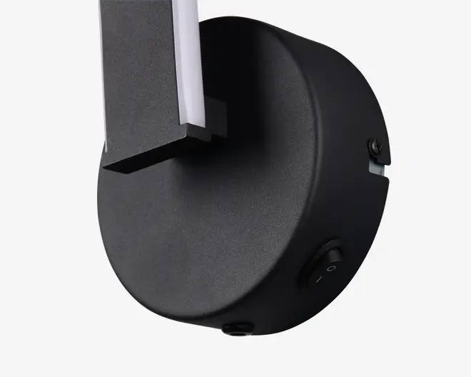 Nærbillede af en vægmonteret lampefod i mat sort med en synlig tænd/sluk vippekontakt på siden. Lampen har et moderne, minimalistisk design med rene linjer og en rektangulær arm, der strækker sig ud fra den runde base. Armen har en hvid stribe, sandsynligvis til LED-belysning. Overfladen er glat, og monteringsbeslaget er diskret integreret.