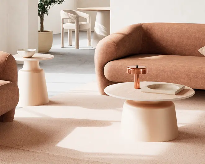 Rundt sofabord med en glat, lysebeige fod og en matchende cirkulær bordplade. Bordet har et minimalistisk design med en bred, solid base og en sømløs overflade. På bordpladen står en moderne kobberlysestage og et lavt, rundt fad. Bordet står på et blødt, neutralt tæppe, der matcher den buede sofa og lænestol i rummet.