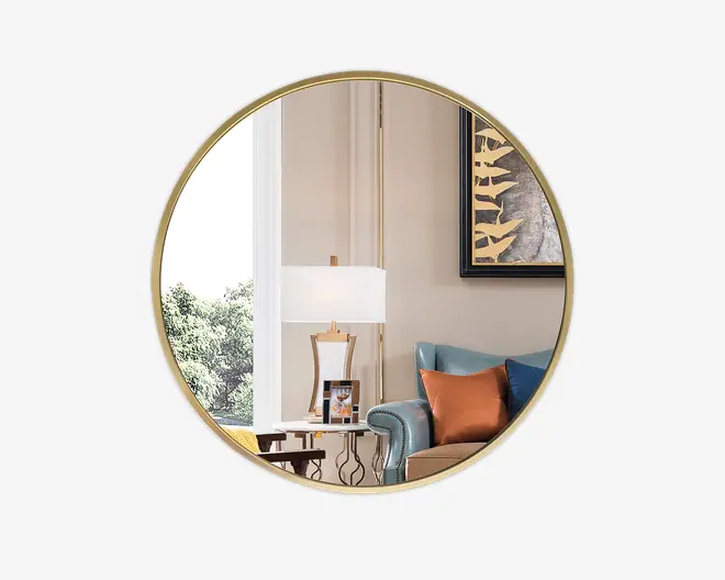 Rundt vægspejl med en tynd, guldfarvet metalramme. Spejlet reflekterer en moderne stue med en blå polstret lænestol, orange og blå pyntepuder, et sidebord med en lampe og en fotoramme samt en del af et vindue med grønt udenfor. Spejlets ramme er slank og minimalistisk, hvilket giver et elegant præg til indretningen.