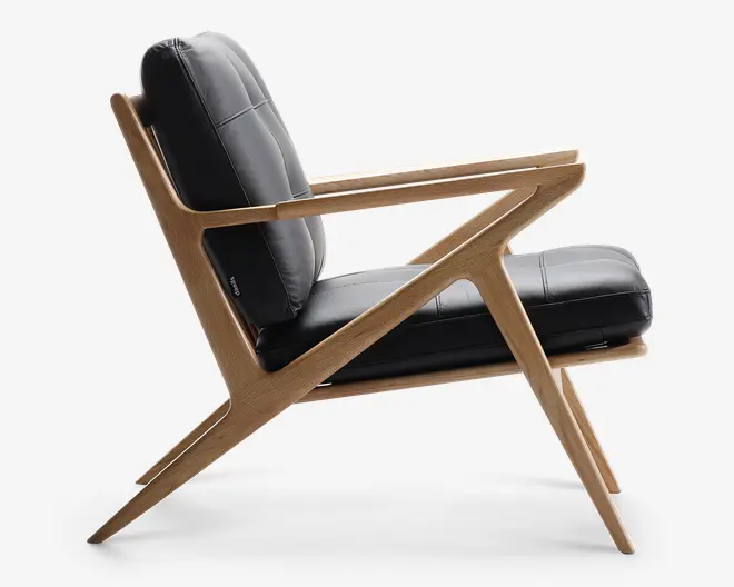 Sideview af en moderne loungestol med en markant, vinklet træramme i naturfinish. Stolen har tykke, sorte læderhynder på både sæde og ryg, hvilket giver et komfortabelt og stilfuldt udtryk. Armlænene er integreret i rammen, og benene er skråtstillede for stabilitet og et moderne udseende.