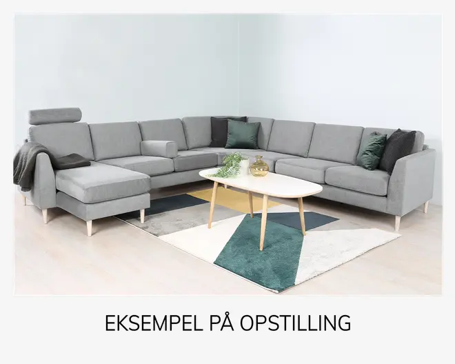 Moderne L-formet hjørnesofa betrukket med lysegråt stof, med rene linjer og slanke, lyse træben. Sofaen har en chaiselong-del, justerbar nakkestøtte og en matchende rektangulær pude. Der er flere dekorative pyntepuder i mørkegrøn og sort på sofaen samt et mørkt tæppe lagt over armlænet. Foran sofaen står et hvidt ovalt sofabord med lyse træben, placeret på et geometrisk mønstret tæppe i nuancer af teal, grå og hvid. Udtrykket er moderne og indbydende.