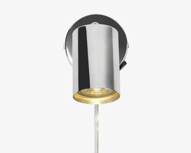 Væghængt lampe med cylindrisk form og blank kromfinish. Lampen har en rund base og en nedadrettet lyskilde med en klar linse i bunden. Designet er moderne og minimalistisk, velegnet til nutidige indretninger.