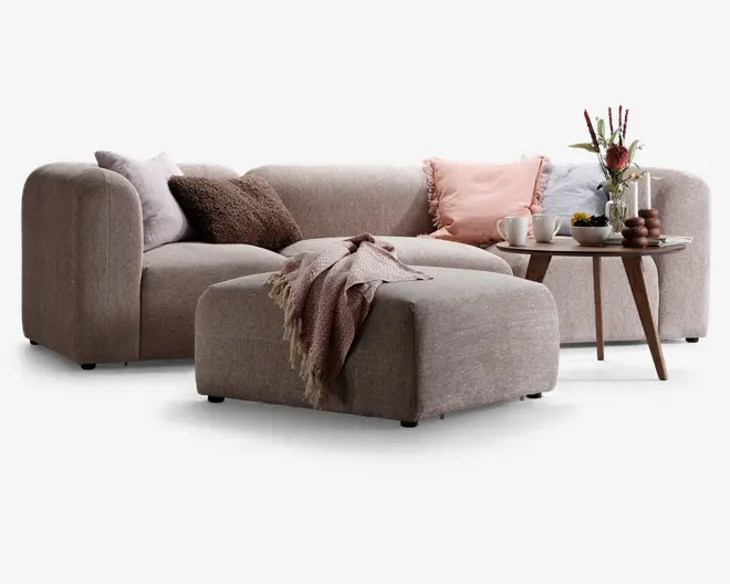 En moderne, L-formet sofa med afrundede kanter og blødt, lysebeige stofbetræk. Sofaen er pyntet med flere dekorative puder i nuancer af pink, brun og grå, hvilket giver et hyggeligt udtryk. Foran sofaen står en matchende puf med et tekstureret, frynset tæppe lagt over. Ved siden af står et rundt sofabord i træ med en vase med blomster, stearinlys og et fad med kopper og en skål, hvilket skaber en varm og indbydende stemning.