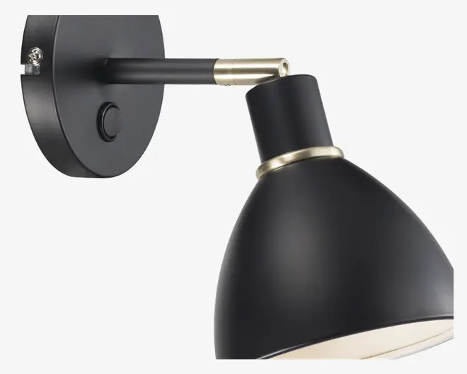 Væghængt lampe i mat sort med konisk skærm, messingfarvet ring ved skærmens base og tilsvarende messingdetalje på armen. Lampen har en rund vægplade med synlig tænd/sluk-knap og et stilrent, moderne design.