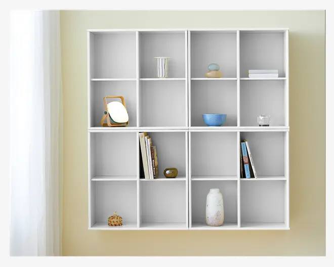 Et vægmonteret reolsystem med et gitter af tolv kvadratiske rum. Hylderne er hvide og har et minimalistisk design. Nogle af rummene indeholder dekorative genstande såsom et lille spejl med træramme, en stribet kop, stablede sten, en blå skål, et glas, en vase, en lille skulptur og nogle bøger placeret lodret og let skråt. Det samlede udtryk er rent og organiseret med god plads til opbevaring eller udstilling.