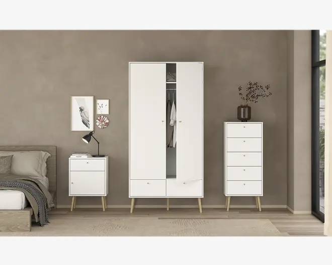 Billedet viser et sæt moderne soveværelsesmøbler med minimalistisk design. Møblerne er hvide med lyse træben og består af et højt garderobeskab med to låger og to skuffer nederst, en kommode med fem skuffer og et natbord med to skuffer. Garderobeskabet har en låge let åben, så man kan se hængende tøj indeni. Natbordet er dekoreret med en lampe og bøger, mens kommoden har en dekorativ vase med grene. Møblerne står op ad en neutral taupefarvet væg, og rummet har en hyggelig, moderne atmosfære.