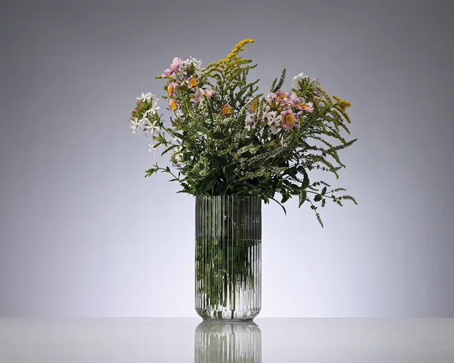 Høj, klar rillet glasvase med afrundet bund, fyldt med en løs buket af blandede vilde blomster i rosa, hvid, gul og grøn.