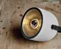 Moderne lampe med cylindrisk krop i beton og en glat, mat overflade. Indersiden har en guldfarvet, reflekterende overflade, der forstærker lyset, samt en synlig klar pære i midten. Lampen har en sort metalring omkring åbningen og en sideplaceret sort metalarm til montering eller justering.
