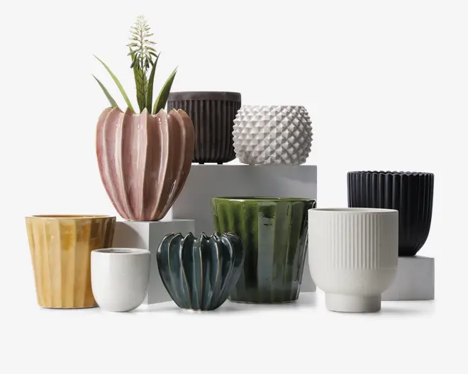 En samling keramiske urtepotter i forskellige former, størrelser og farver. Potterne har forskellige teksturer, herunder riflede, fløjlsbløde og piggede overflader. Farverne spænder fra pastelrosa, gul og hvid til dyb grøn, mørkeblå og sort. Nogle potter har blank finish, mens andre er matte. Én potte indeholder en plante med lange grønne blade og en hvid blomst.