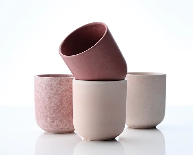 Sæt med fire minimalistiske, cylindriske kopper i bløde pastelfarver af rosa og beige. Hver kop har en glat, mat overflade med diskrete variationer i tekstur og farve, hvilket giver et håndlavet udtryk. Kopperne har lige sider med en let afrundet bund og ingen hank, hvilket gør dem velegnede til både varme og kolde drikke. Designet er moderne og elegant, perfekt til et nutidigt borddækning.