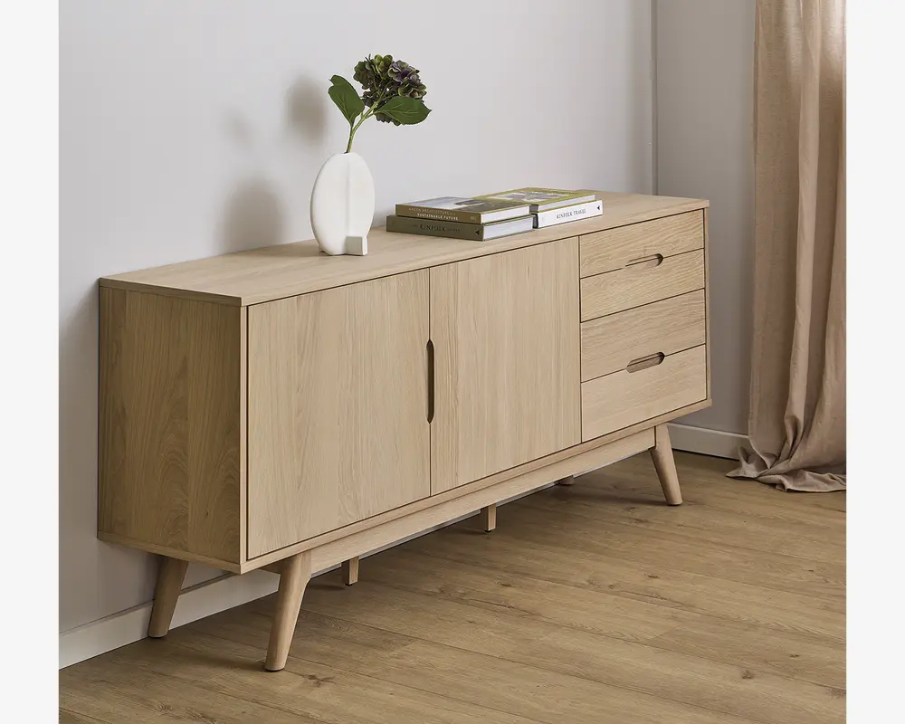 Lys egetræsskænk med minimalistisk design, to centrale låger med indbyggede greb og tre skuffer til højre. Skænken står på skråtstillede, koniske ben og har en glat, naturlig træfinish. Toppen er dekoreret med en hvid vase med grønne blade og to stabelde bøger.