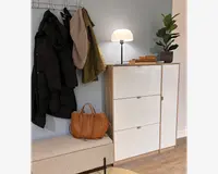 Et moderne entréarrangement med et opbevaringsskab i hvid og træfinish med tre rum og slanke metalhåndtag. Oven på skabet står en lille bordlampe med rund, hvid skærm, en stak trækasser og en potteplante med brede grønne blade. Til venstre hænger en vægmonteret knagerække med en sort dunjakke, en grøn frakke og et beige strikket halstørklæde. Under knagerækken står en polstret bænk i et lyst, tekstureret stof, hvorpå der ligger en lysebrun læderhåndtaske. Gulvet er i træ, og væggen er malet i en blød blågrå farve.
