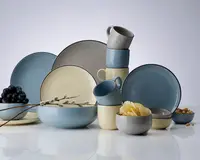 Et sæt matchende keramikservice bestående af tallerkener, skåle og krus i bløde pastelfarver som blå, creme og grå. Delene har en glat, mat overflade med afrundede kanter og et minimalistisk design. Sættet indeholder store middagstallerkener, mindre sidetallerkener, dybe skåle og stabelbare krus, alle med et moderne og stilrent udtryk. Nogle af skålene er fyldt med madvarer som vindruer, chips og nødder for at vise anvendelsen. Stilen er moderne og elegant, velegnet til både hverdag og fest.