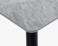Nærbillede af et bordhjørne med en glat, rektangulær bordplade med afrundede kanter. Bordpladen har et lysegråt marmorlignende mønster og en let skrå kant. Bordbenet er cylindrisk og sort, hvilket giver en moderne kontrast til den lyse overflade.