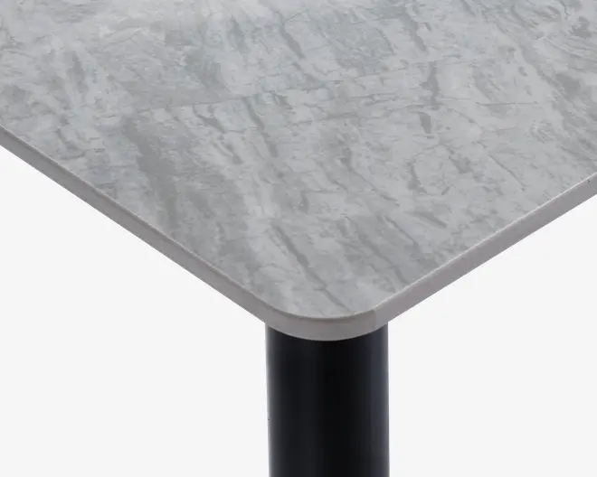Nærbillede af et bordhjørne med en glat, rektangulær bordplade med afrundede kanter. Bordpladen har et lysegråt marmorlignende mønster og en let skrå kant. Bordbenet er cylindrisk og sort, hvilket giver en moderne kontrast til den lyse overflade.