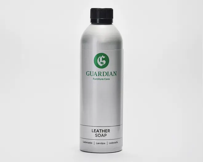 En høj, cylindrisk sølvfarvet flaske med sort skruelåg. Flasken har et grønt 'G'-logo og mærket 'Guardian Furniture Care' trykt i grønt. Nederst på etiketten står der 'Leather Soap' med fed sort skrift, og derunder er der oversættelser på flere sprog. Designet er stilrent og moderne med en metallisk overflade.