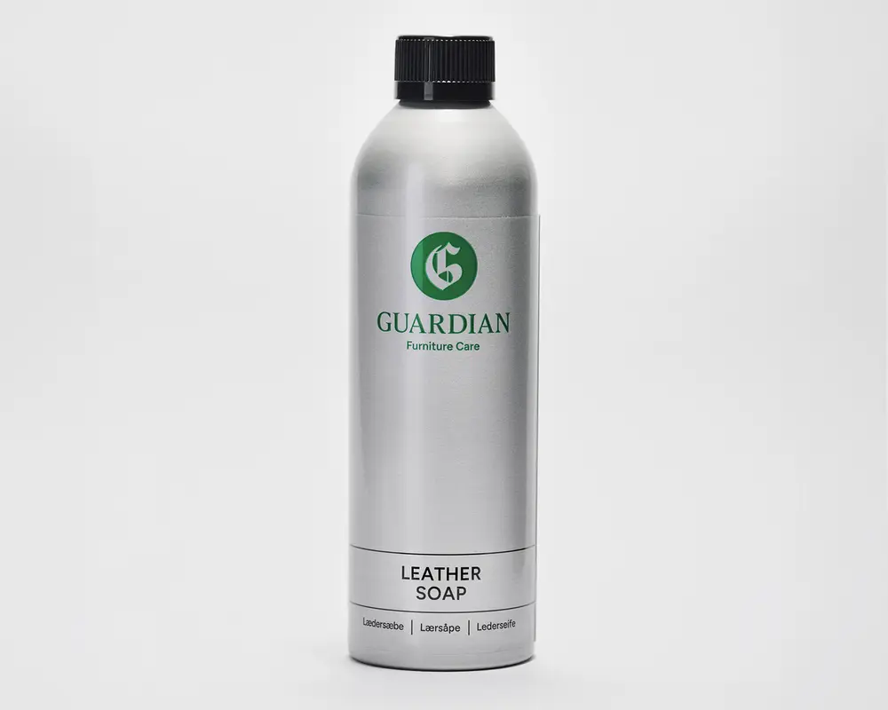En høj, cylindrisk sølvfarvet flaske med sort skruelåg. Flasken har et grønt 'G'-logo og mærket 'Guardian Furniture Care' trykt i grønt. Nederst på etiketten står der 'Leather Soap' med fed sort skrift, og derunder er der oversættelser på flere sprog. Designet er stilrent og moderne med en metallisk overflade.