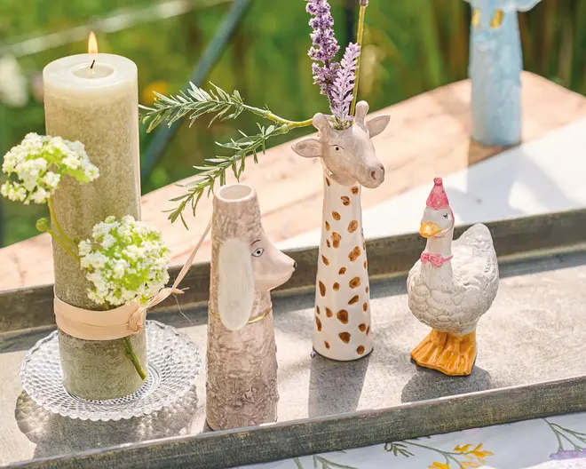 Billedet viser en dekorativ opstilling med tre keramiske dyrefigurer: en høj girafvase med brune pletter og lavendelstilke, en hundevase med tekstureret overflade og gyldent halsbånd samt en andefigur med lyserød festhat og orange fødder. Ved siden af står et grønt bloklys omviklet med beige bånd og pyntet med hvide blomster. Alle genstande er placeret på et rektangulært metalfad, der står udendørs på et træbord.