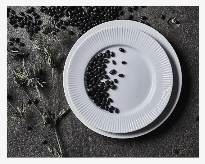 To stablede hvide tallerkener med rillet, tekstureret kant. Tallerkenerne er runde og har en blank overflade. Den øverste tallerken har spredte sorte bønner på sig, og baggrunden er en ru, mørk stenoverflade med tørrede plantestængler og flere sorte bønner spredt omkring.