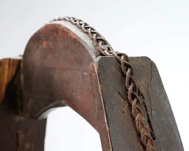 Nærbillede af et mørkt, vejrslidt træstykke med en buet udskæring og en ældet metalkæde lagt langs overkanten; overfladen har tydelige slidmærker, ridser og en rustfarvet patina.