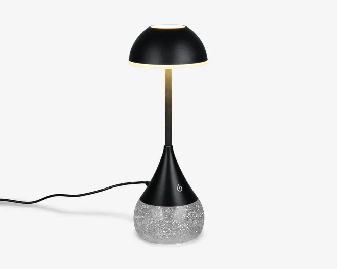 Moderne bordlampe med en elegant, sort kuppelformet skærm og en slank, metallisk stang. Basen er konisk og sort med en touch-følsom tænd/sluk-knap og står på en klar, struktureret del fyldt med glitrende glimmer for et dekorativt udtryk. Lampen har en synlig ledning til strøm.