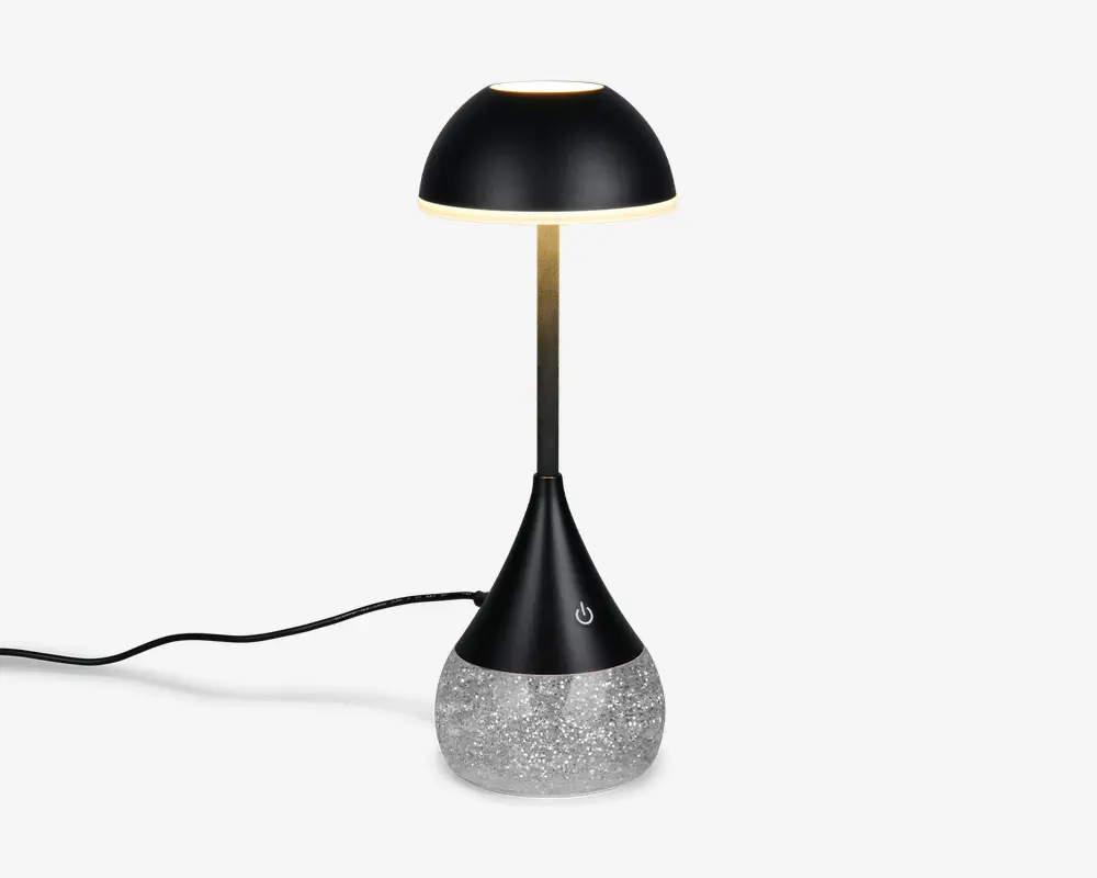 Moderne bordlampe med en elegant, sort kuppelformet skærm og en slank, metallisk stang. Basen er konisk og sort med en touch-følsom tænd/sluk-knap og står på en klar, struktureret del fyldt med glitrende glimmer for et dekorativt udtryk. Lampen har en synlig ledning til strøm.