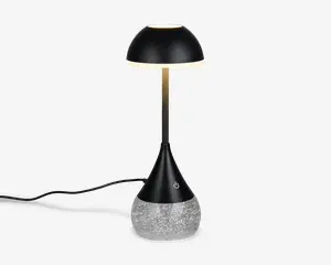 Moderne bordlampe med en elegant, sort kuppelformet skærm og en slank, metallisk stang. Basen er konisk og sort med en touch-følsom tænd/sluk-knap og står på en klar, struktureret del fyldt med glitrende glimmer for et dekorativt udtryk. Lampen har en synlig ledning til strøm.