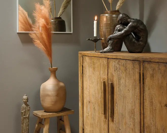 En rustik boligindretning med en mat beige keramisk vase fyldt med høje orange pampasgræsstrå på en lille træskammel ved siden af et slidt træskab, som er pyntet med en mørk skulpturfigur, et tændt stearinlys og en металisk vase; en lille dekorativ figur står på gulvet.