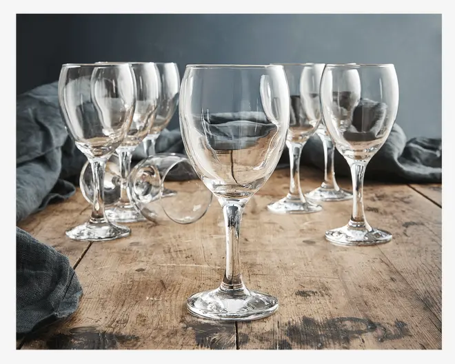 Sæt med seks klare glasbægre med klassisk rund skål og solid stilk, arrangeret på en rustik træoverflade. Glassene har en glat, poleret finish og en let udadbuet kant, velegnet til servering af vin eller andre drikkevarer. Ét glas ligger på siden, hvilket viser glassets tykkelse og klarhed.