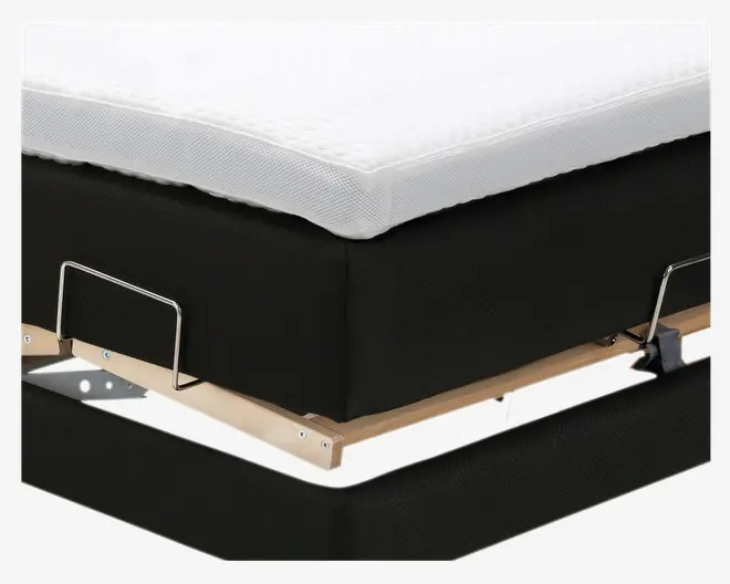 Nærbillede af en sengebund med sort stofbetræk, der har en solid ramme af trælameller og metalstøttebøjler. Øverst ses en hvid, tekstureret madras eller topmadras med åndbart mesh-stof. Konstruktionen virker robust med synlige metalbeslag og forstærket hjørne for ekstra stabilitet.