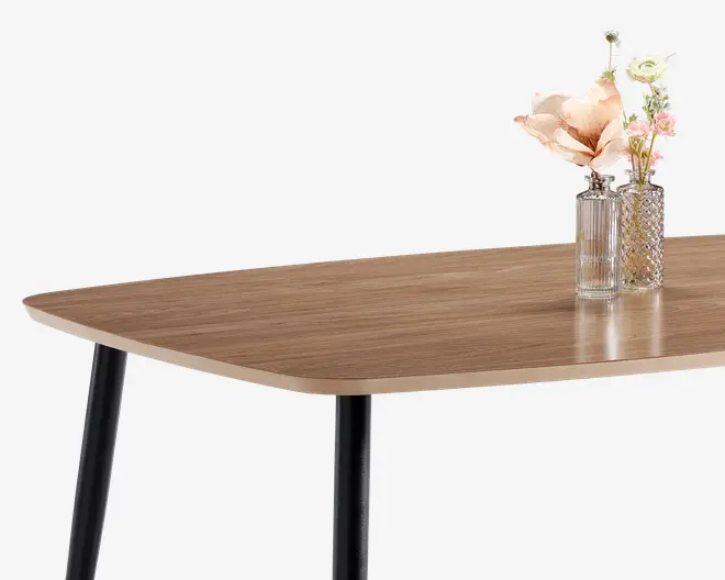 Rektangulært bord med en glat, lys træfinish og afrundede hjørner. Bordet har solide sorte metalben og er dekoreret med to glasvaser på toppen, den ene med et mønstret design og den anden med lodrette riller, begge med blomster i pastelfarver.