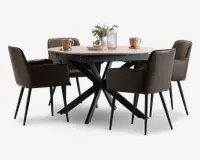 Et rundt spisebord med en lys træbordplade og en moderne sort metalbase med krydsende ben. Bordet er omgivet af fire polstrede stole i mørkebrun farve, hver med armlæn og slanke sorte ben. På bordpladen står der to lyserøde krus på underkopper, en lyserød rillet vase med grønne grene og en dekorativ sølvskål, hvilket skaber en stilfuld og indbydende atmosfære.