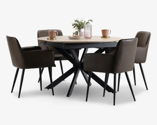 Et rundt spisebord med en lys træbordplade og en moderne sort metalbase med krydsende ben. Bordet er omgivet af fire polstrede stole i mørkebrun farve, hver med armlæn og slanke sorte ben. På bordpladen står der to lyserøde krus på underkopper, en lyserød rillet vase med grønne grene og en dekorativ sølvskål, hvilket skaber en stilfuld og indbydende atmosfære.