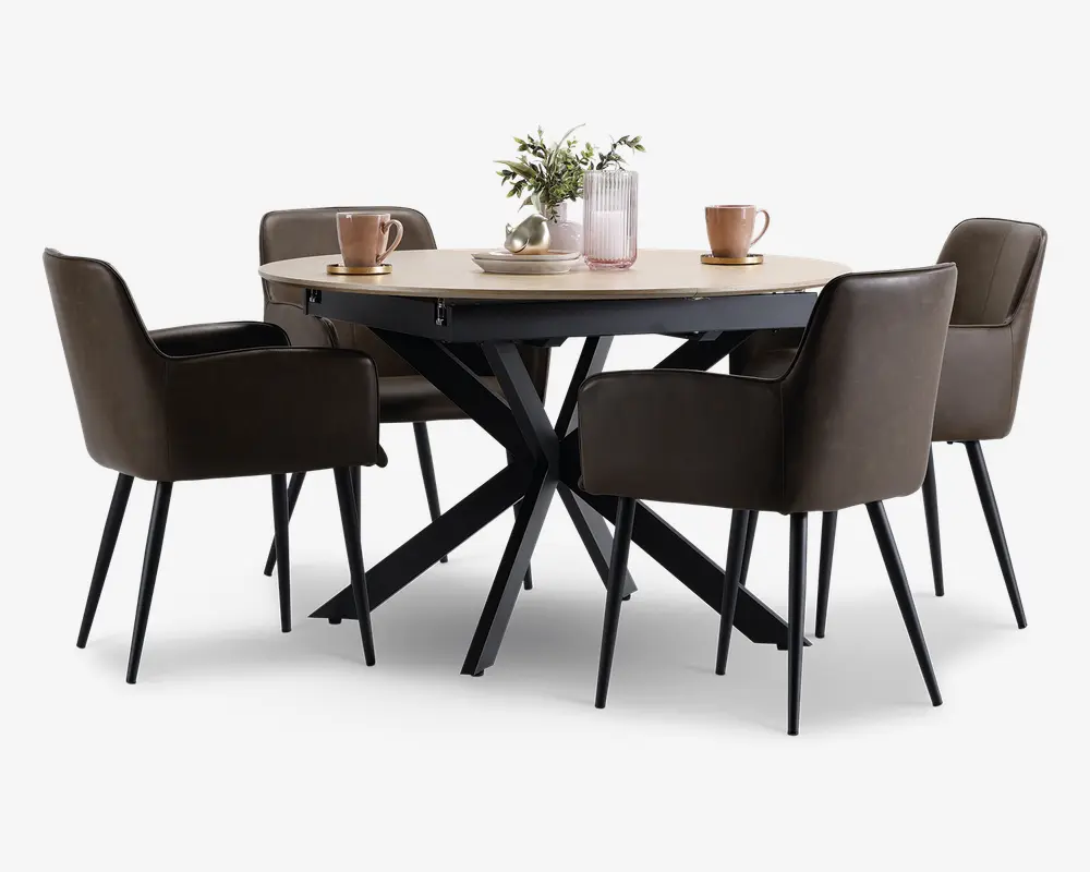 Et rundt spisebord med en lys træbordplade og en moderne sort metalbase med krydsende ben. Bordet er omgivet af fire polstrede stole i mørkebrun farve, hver med armlæn og slanke sorte ben. På bordpladen står der to lyserøde krus på underkopper, en lyserød rillet vase med grønne grene og en dekorativ sølvskål, hvilket skaber en stilfuld og indbydende atmosfære.