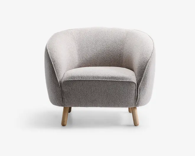 En moderne lænestol med rundet ryg og sider, polstret med lysegråt tekstureret stof. Sædet er bredt og polstret for komfort. Stolen står på fire korte, skråtstillede træben med naturfinish, hvilket giver et moderne og hyggeligt udtryk.