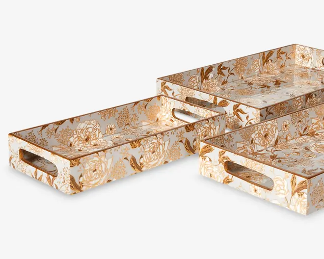 Sæt med tre rektangulære bakker med høje kanter og udskårne håndtag i de korte ender. Hver bakke har en blank overflade med et detaljeret guld- og hvidt blomstermønster, hvilket giver dem et elegant og dekorativt udseende. Bakkerne er stablet i hinanden og viser deres forskellige størrelser.